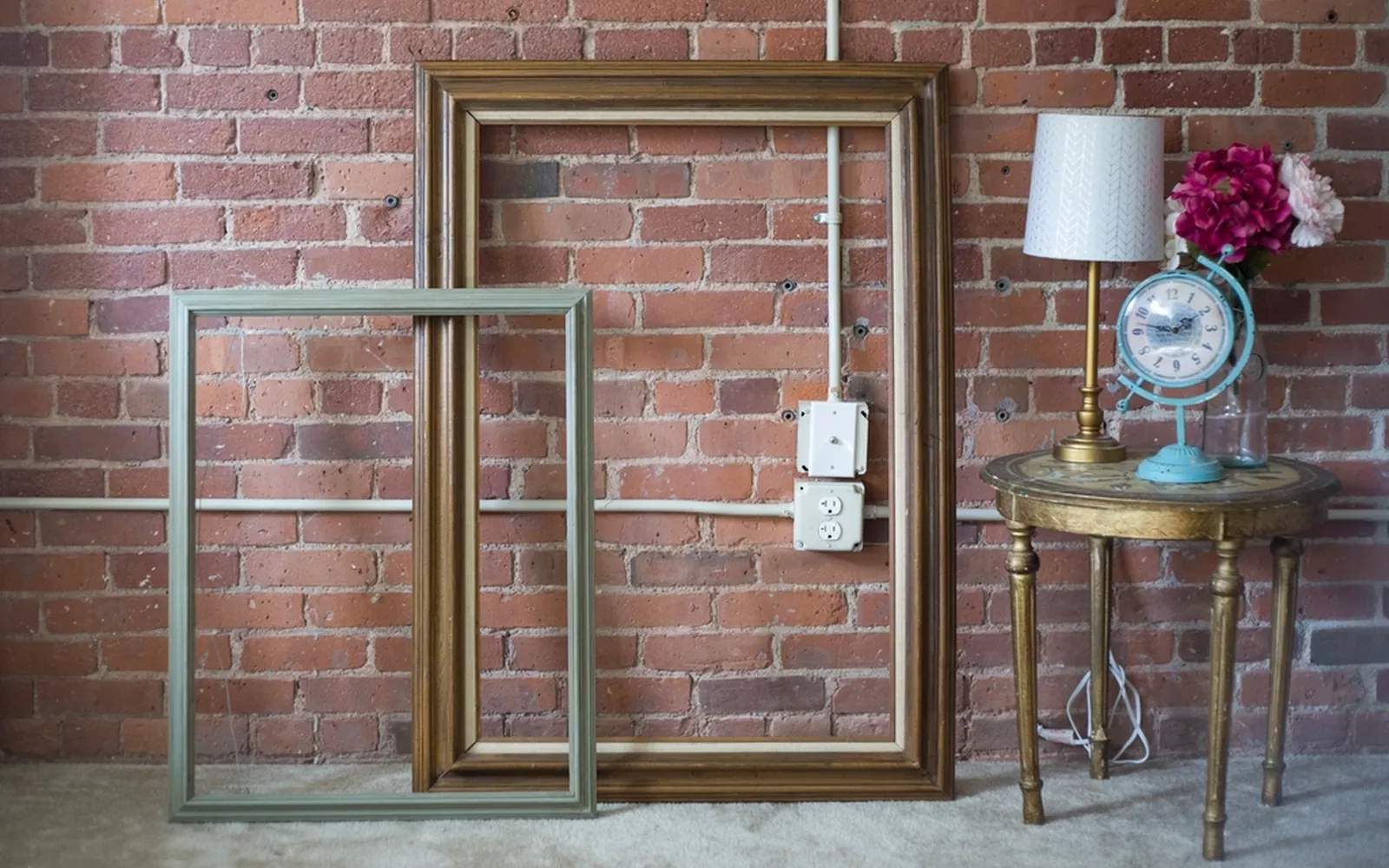 Empty wooden picture frames leaning against a brick wall next to a lamp and flowers