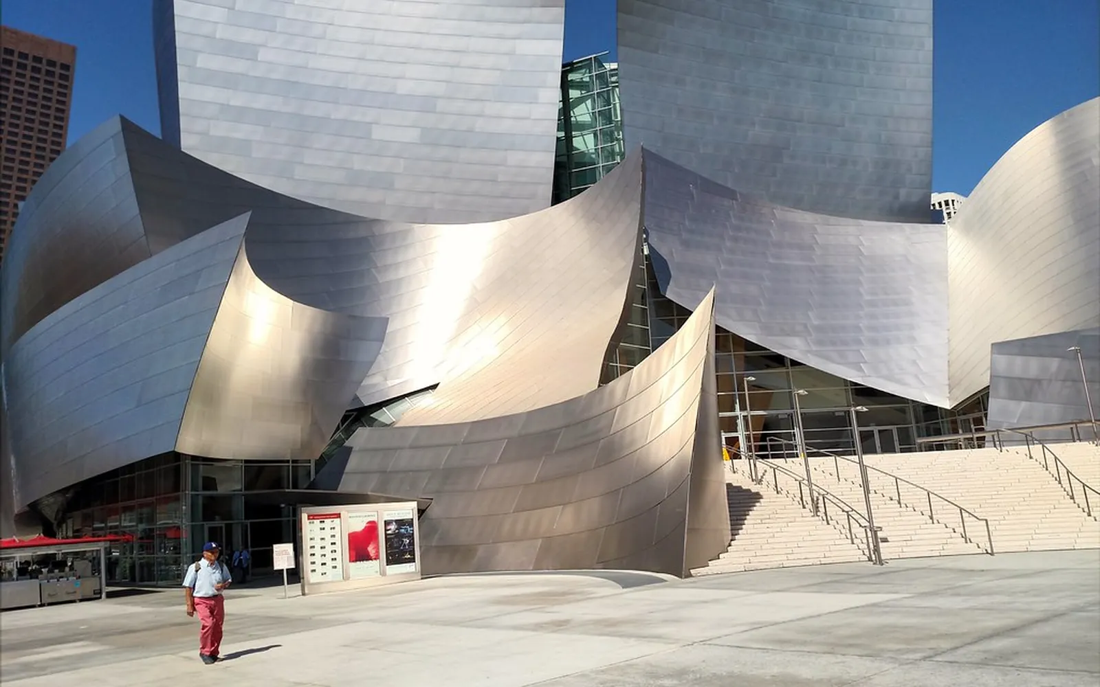 Curved stainless steel panels of the Walt Disney Concert Hall in Los Angeles under a clear blue sky, with steps and a pedestrian in front